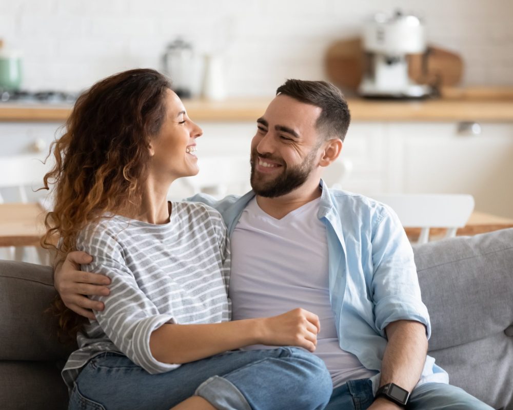 Happy young couple of man and woman embracing look at each other sitting on couch in living room. Attractive smiling husband and wife laughing having fun free time together.